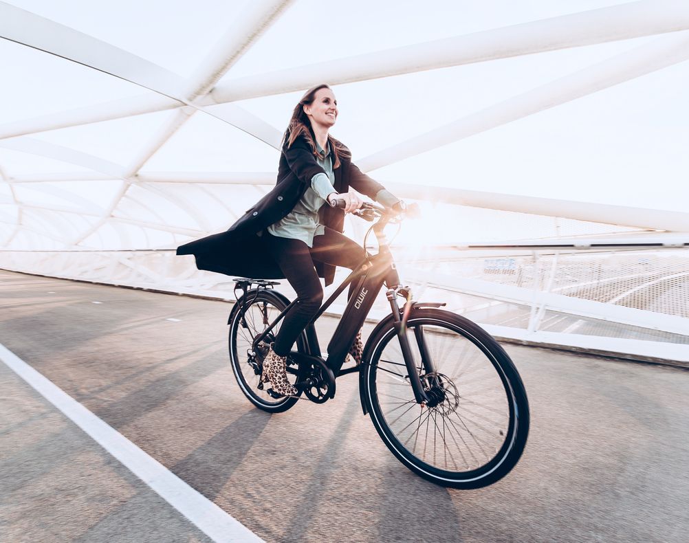 Frau in Mantel fährt Fahrrad auf einer modernen Brücke bei Sonnenlicht