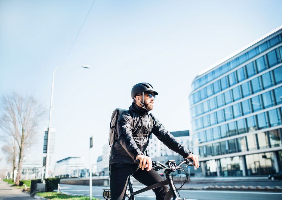 Person mit Helm und Rucksack fährt auf einem E-Bike in urbaner Umgebung mit modernen Bürogebäuden