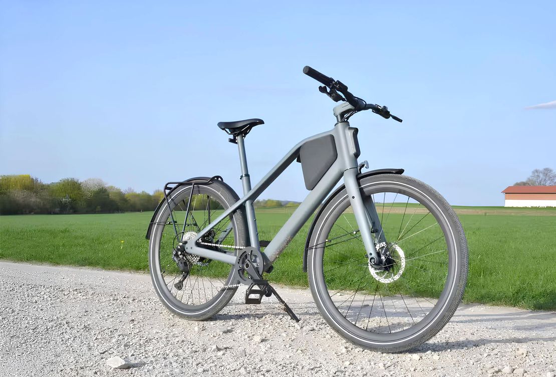 Elektrofahrrad mit grauem Rahmen und breiten Reifen steht auf einem Schotterweg vor grüner Wiese und blauem Himmel