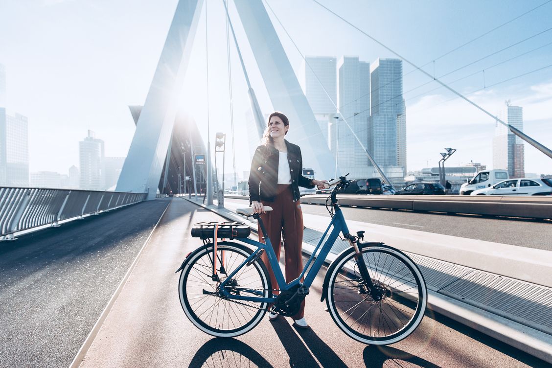 Frau steht mit blauem Fahrrad auf einer Brücke vor einer modernen Stadtlandschaft im Sonnenlicht