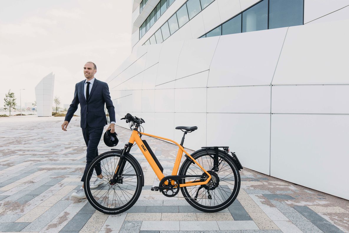 Ein Mann im Anzug geht neben einem orangefarbenen Fahrrad vor einem modernen Gebäude mit weißer Fassade